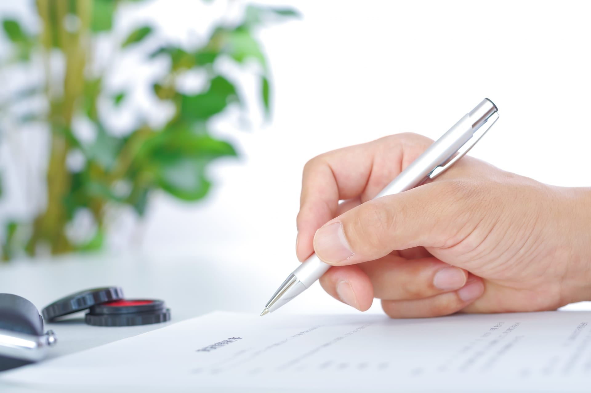 白いデスクの上で、書類にボールペンで記入しようとしている手元のクローズアップ