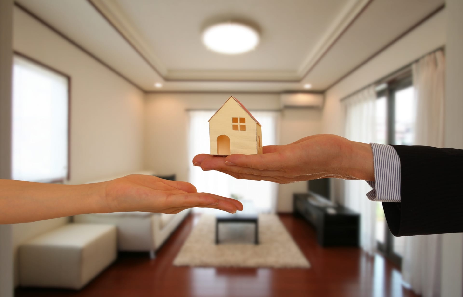 室内のリビングを背景に、手のひらの上で家のミニチュア模型が受け渡されている